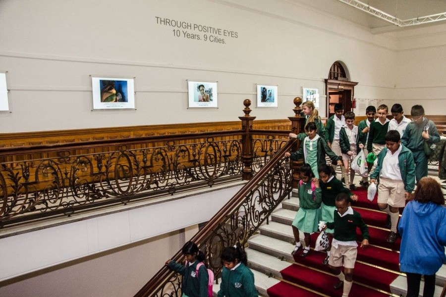 Schoolchildren walking down a staircase past photographs and autobiographical statements by Through Positive Eyes artivists