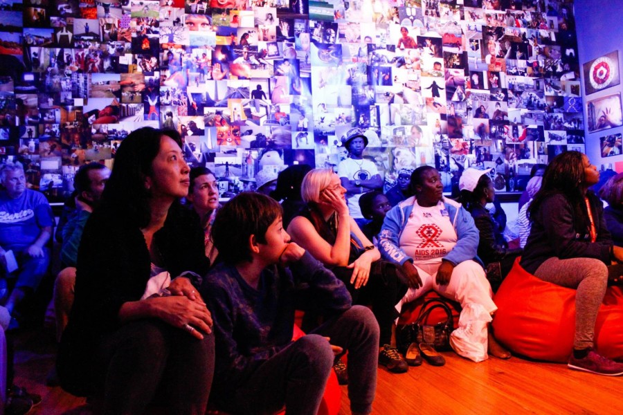 Visitors seated on red beanbag chairs watching a performance in a room wallpapered with Through Positive Eyes photographs
