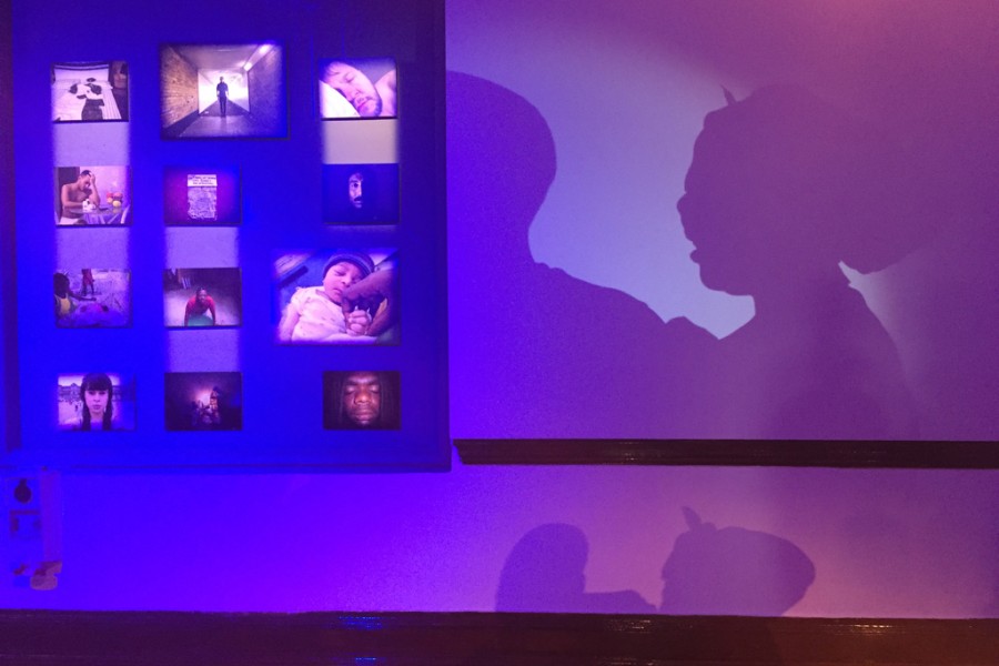 Silhouettes of an artivist against photographs under blue and pink lighting in the Banishing Stigma Room